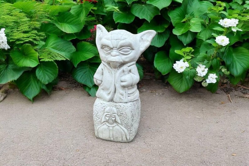 May include: A light gray Yoda statue with a detailed face and robe, standing on a base with a Darth Vader image. The statue is outdoors, surrounded by green foliage and white flowers, creating a garden decor theme.