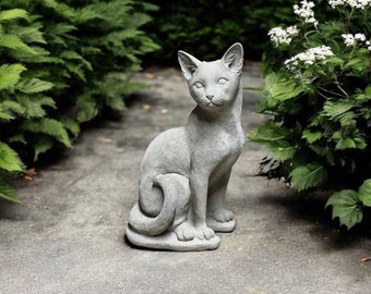 May include: A gray concrete cat statue sitting on a gray stone surface. The cat is looking directly at the camera with its tail curled around its body.