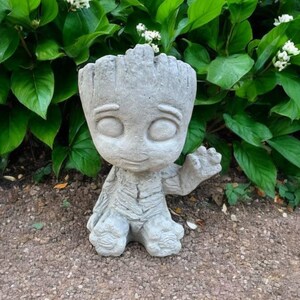 May include: A gray concrete garden statue of a character from the movie Guardians of the Galaxy. The statue is of Groot, a tree-like humanoid, with a friendly expression and a raised hand. The statue is sitting on a bed of gravel.