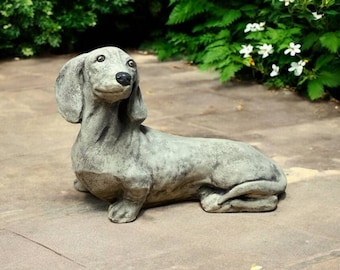 May include: A gray concrete dachshund statue in a relaxed pose, with its head lifted and ears flopped. The dog sculpture has a detailed texture and is set on a stone patio, surrounded by greenery and white flowers.