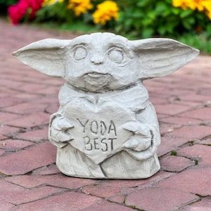 May include: A gray concrete statue of a character holding a heart-shaped sign. The sign reads "YODA BEST". The statue has large ears and a detailed face. The statue is outdoors on a brick surface.