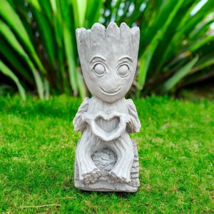 May include: A light gray, tree-like humanoid planter with a textured bark surface. The figure has large, round eyes and is sitting on a log-shaped base. Its hands are positioned in front of its chest, forming a heart shape with its fingers.
