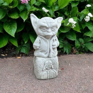 May include: A gray concrete Yoda statue with large ears, a robe, and a staff. The statue is on a base with a carved image of Darth Vader. The statue is outdoors, surrounded by green foliage and flowers.