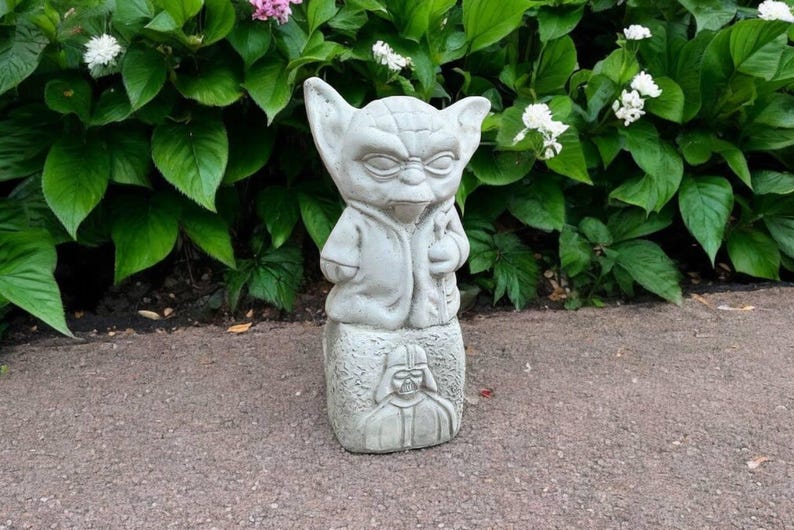May include: A gray concrete Yoda statue with large ears, a robe, and a staff. The statue is on a base with a carved image of Darth Vader. The statue is outdoors, surrounded by green foliage and flowers.