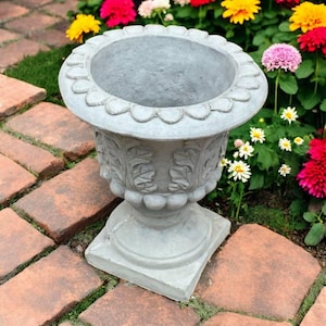 May include: A gray, ornate garden urn with a scalloped rim and detailed floral carvings. The urn is set on a square base and surrounded by colorful flowers and brick pavers, perfect for outdoor decor.