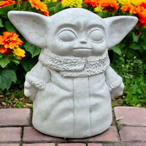 May include: A light gray concrete statue of a character with large ears, large eyes, and a small mouth. The statue is wearing a scarf and a coat. The statue is standing on a brick surface, with colorful flowers in the background.
