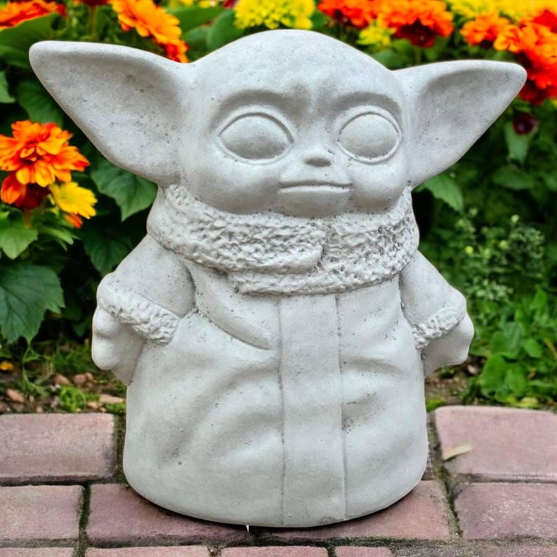 May include: A light gray concrete statue of a character with large ears, large eyes, and a small mouth. The statue is wearing a scarf and a coat. The statue is standing on a brick surface, with colorful flowers in the background.