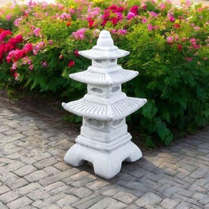 May include: A white, three-tiered pagoda-style garden statue with a pointed top. The statue is made of stone and sits on a brick pathway, with a backdrop of vibrant pink and green flowering bushes.