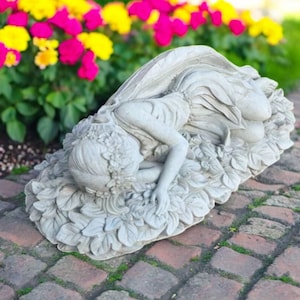 May include: A grey stone fairy statue, lying on a bed of leaves and flowers. The fairy has detailed wings and wears a floral crown. The statue is outdoors, with a backdrop of pink and yellow flowers.