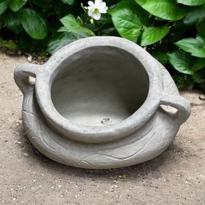 May include: A gray ceramic planter with a wide opening and two handles. The planter has a textured surface and is sitting on a gravel path.