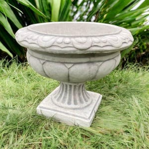 May include: A gray, ornate concrete planter with a wide, decorative rim. The planter has a bulbous body and sits on a square base. The design includes detailed floral patterns. The planter is outdoors on a bed of green grass.