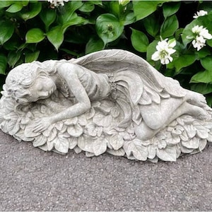 May include: A gray concrete garden statue of a fairy lying on a bed of leaves. The fairy has wings and is wearing a flowing dress.
