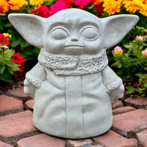 May include: A light gray concrete statue of a character with large ears and a hooded coat. The statue has large eyes and a neutral expression. The statue is on a brick surface with colorful flowers in the background. The statue is a decorative garden ornament.
