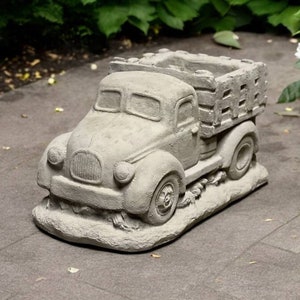 May include: A gray concrete planter shaped like a vintage pickup truck. The truck bed is open and has wooden slats. The truck is sitting on a gray concrete surface.