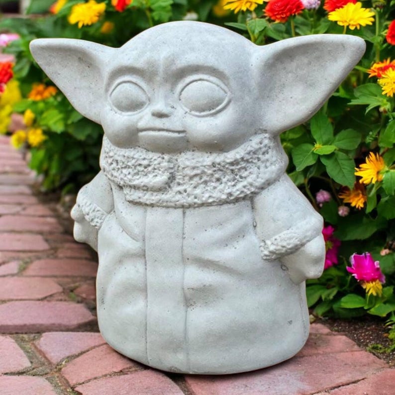 May include: A light gray statue of a character with large ears, large eyes, and a textured collar. The statue is standing in front of a colorful flower bed. The statue is a decorative garden ornament.