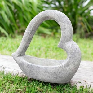 May include: A gray, heart-shaped planter made of stone. The planter has a textured surface and a wide opening for plants. The heart shape is formed by a curved handle that rises from the base. The planter is sitting on a wooden surface.