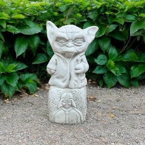 May include: A gray concrete Yoda statue with a Darth Vader base stands in front of green foliage. The Yoda figure has large ears and is wearing a robe. The base features a relief of Darth Vader's helmet.