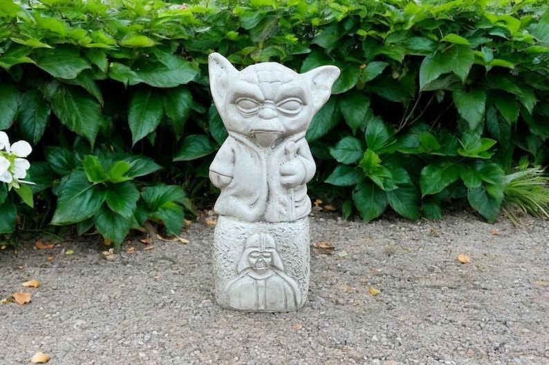 May include: A gray concrete Yoda statue with a Darth Vader base stands in front of green foliage. The Yoda figure has large ears and is wearing a robe. The base features a relief of Darth Vader's helmet.