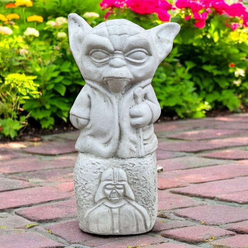 May include: A gray concrete garden statue featuring Yoda from Star Wars. The statue has large ears and is holding a staff. Below Yoda is an image of Darth Vader. The statue is set on a brick walkway with flowers in the background.