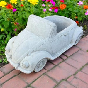 May include: A light gray concrete planter shaped like a classic convertible car. The planter has a large open space for planting flowers or other greenery. The car planter is sitting on a brick surface, with colorful flowers in the background.
