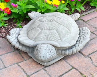 May include: A gray concrete turtle statue with detailed shell and flipper designs. The turtle is positioned on a brick surface, with colorful flowers in the background. A decorative garden ornament.