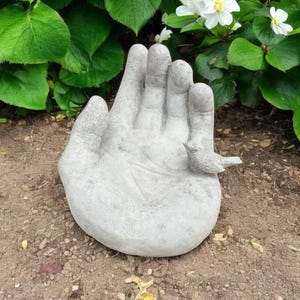 以下が含まれることがあります： 手のひらに小さな鳥がとまっている灰色のコンクリート製のハンド彫刻。手は開いており、鳥は右を向いています。彫刻は茶色の土の上に置かれています。