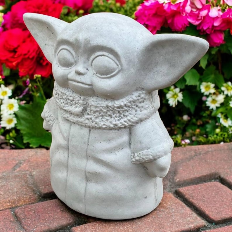 May include: A gray concrete statue of a character with large ears and a textured scarf. The statue is outdoors, set against a backdrop of vibrant pink and white flowers, and a red brick surface.