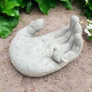 以下が含まれることがあります： 手のひらに鳥がとまっているコンクリート製のハンド彫刻。手は砂利のベッドの上に置かれています