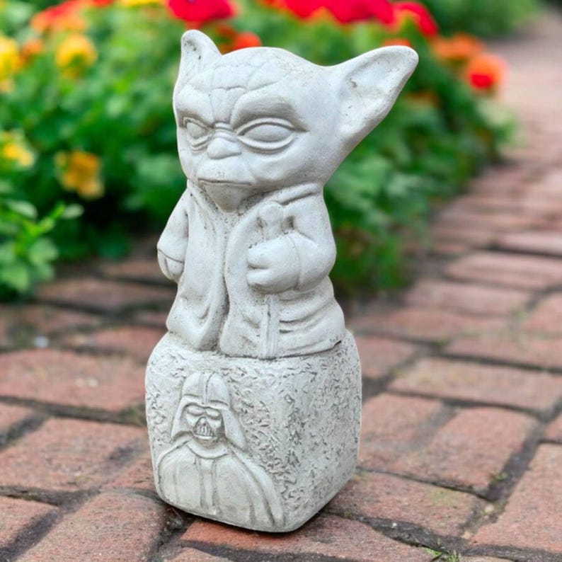 May include: A light gray Yoda statue with large ears and a robe, standing on a base with a Darth Vader image. The statue is made of a stone-like material and is placed outdoors near a brick walkway and colorful flowers.