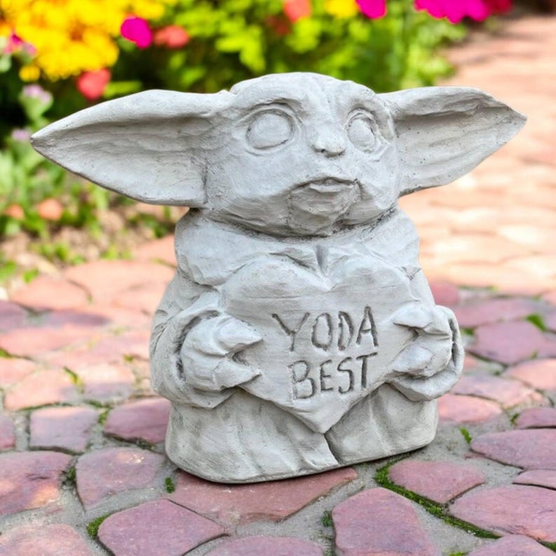 May include: A gray concrete statue of a character holding a heart-shaped sign that reads "YODA BEST." The statue has large ears and a detailed face. The statue is a garden ornament.