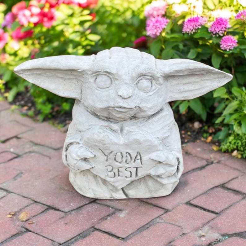 May include: A light gray statue of a character with large ears, holding a heart-shaped sign that reads "YODA BEST". The statue is outdoors, surrounded by greenery and flowers, and is made of a stone-like material.
