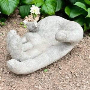 以下が含まれることがあります： 手のひらに鳥がとまっている灰色のコンクリート製のハンド彫刻。手は開いていて、何かを提供しているように見える。