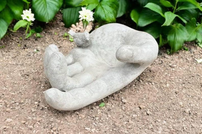 以下が含まれることがあります： 手のひらに鳥がとまっている灰色のコンクリート製のハンド彫刻。手は開いていて、何かを提供しているように見える。