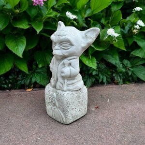 May include: A light gray Yoda statue, standing on a square base with a Darth Vader image. The statue is outdoors, surrounded by green foliage and flowers. The Yoda figure is holding a staff.