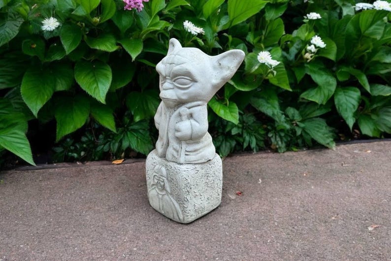 May include: A light gray Yoda statue, standing on a square base with a Darth Vader image. The statue is outdoors, surrounded by green foliage and flowers. The Yoda figure is holding a staff.
