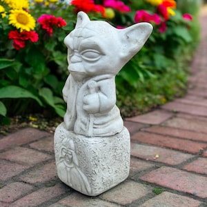 May include: A light gray Yoda statue with a detailed face and robe, standing on a square base with a Darth Vader image. The statue is outdoors, surrounded by colorful flowers and a brick walkway.