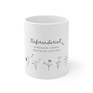 Könnte beinhalten: Weiße Keramiktasse mit schwarzem Text "Referendariat. Gemeinsam lernen. Gemeinsam wachsen." und einer Reihe kleiner schwarzer und brauner Blumen mit Herzen.
