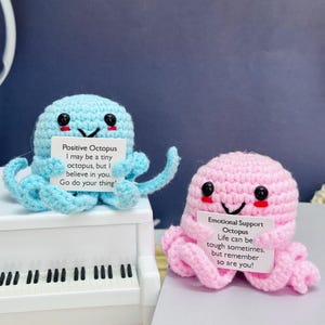 May include: Two crocheted octopus plushies, one blue and one pink, each holding a card with a positive message. The blue octopus says "Positive Octopus I may be a tiny octopus, but I believe in you. Go do your thing!" The pink octopus says "Emotional Support Octopus Life can be tough sometimes, but remember so are you!"