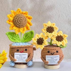 May include: Two handmade crochet sunflower plants in brown pots with smiling faces. The sunflowers are yellow and brown, with green leaves. Each pot has a sign that reads "Emotional Support Plant. Life can be tough sometimes, but remember so are you!"