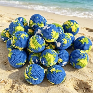 May include: A collection of blue, yellow, and green crocheted balls, each resembling a miniature Earth. The balls are scattered on a sandy beach, with the ocean visible in the background. The handmade spheres feature detailed continent patterns.