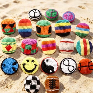 Puede incluir: Una colección de coloridas pelotas y cojines de ganchillo hechos a mano, dispuestos sobre arena. Los diseños incluyen rayas, patrones geométricos y símbolos como una carita sonriente, el yin-yang y una pelota de baloncesto. La paleta de colores es vibrante.