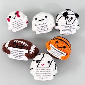 Könnte beinhalten: Eine Sammlung handgehäkelter Sportbälle, darunter Baseball, Golfball, Fußball, Basketball, Volleyball und Fußball. Jeder Ball hat ein lächelndes Gesicht und ein kleines Schild mit einer inspirierenden Botschaft.