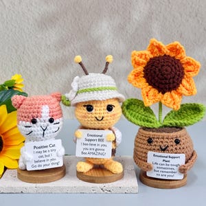 May include: Three crocheted figures: a pink and white cat, a yellow bee, and a sunflower in a pot. Each holds a sign with an inspirational message. The cat's sign reads "Positive Cat." The bee's sign says "Emotional Support BEE." The sunflower's sign reads "Emotional Support Plant."