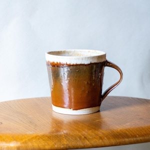 Puede incluir: Una taza de cerámica hecha a mano con un esmalte marrón y blanco. La taza tiene un asa redondeada y una abertura ancha. La taza está sobre una superficie de madera. La taza mide aproximadamente 10 cm de alto.