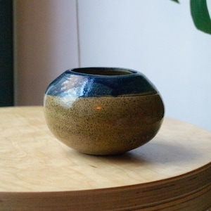 May include: A small, round ceramic vase with a speckled tan body and a dark blue rim. The vase is on a light-coloured wooden surface. The design is simple and modern, suitable for home decor.