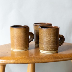 Könnte beinhalten: Drei braune Keramikbecher mit Henkeln stehen auf einem Holztisch. Die Becher haben eine strukturierte Oberfläche und ein helleres Band am Boden. Die Innenseite der Becher ist dunkelbraun.
