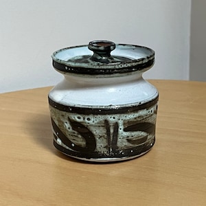 May include: A ceramic jar with a lid. The jar is white with dark brown abstract designs and a dark brown rim. The lid has a small, rounded handle. The jar sits on a light brown wooden surface.