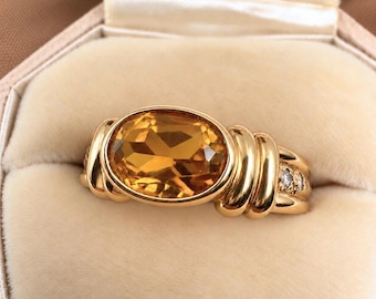 May include: A gold ring featuring a large, oval-cut, yellow citrine gemstone as the centerpiece. The band is crafted with a ribbed, multi-layered design and includes small, clear, round-cut accent stones set along the side of the shank.