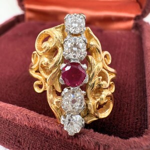 May include: A vintage gold ring with a floral design, featuring three clear diamonds and one red ruby. The ring is set against a burgundy velvet background, showcasing its intricate details and craftsmanship.