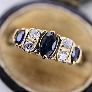 May include: A gold ring featuring a series of gemstones. The ring has a central, oval-cut, dark blue sapphire flanked by smaller, round, clear gemstones and additional dark blue sapphires. The ring is set against a neutral background.